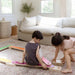 Two children playing on a colorful wooden balance beam in a living room.