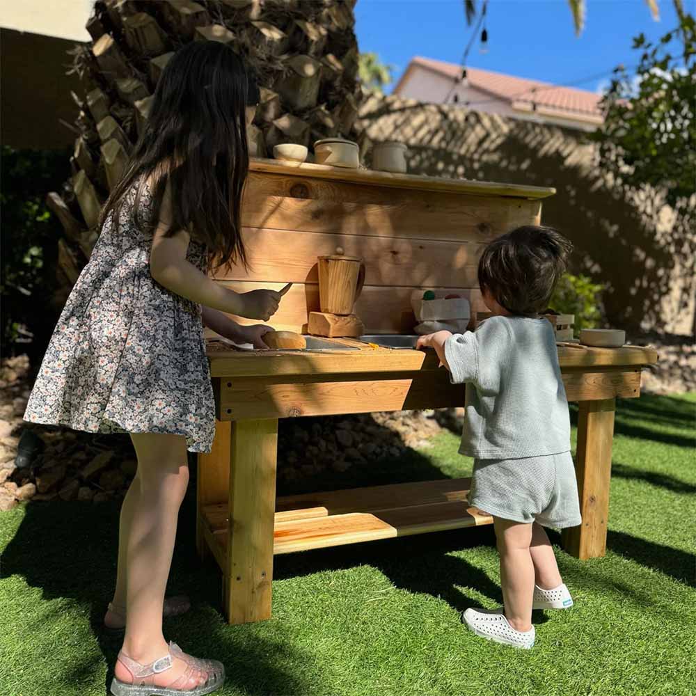 Two children playing at a wooden outdoor mud kitchen with a tree and house in the background.