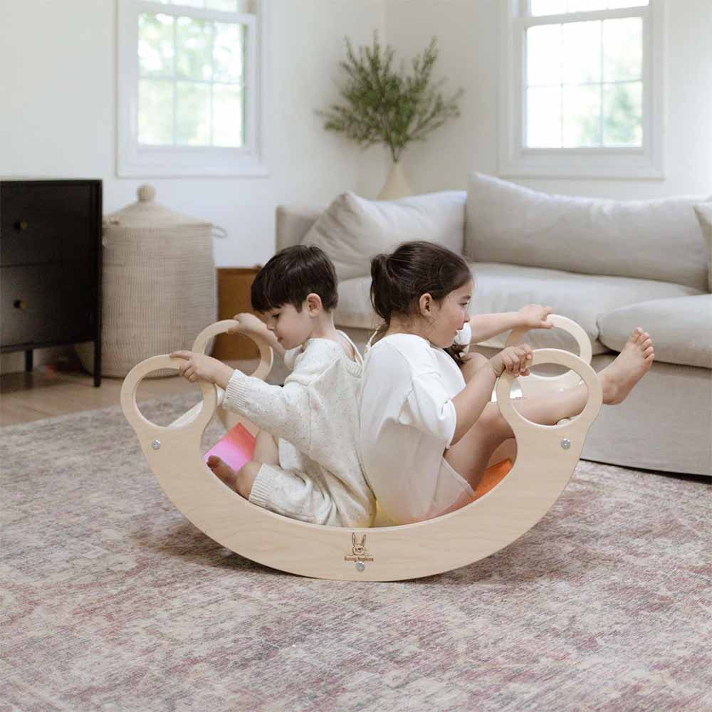 Two children playing on a wooden rocking toy in a living room.