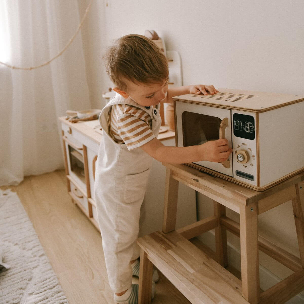 Child playing with a wooden toy microwave in a room with white curtains.