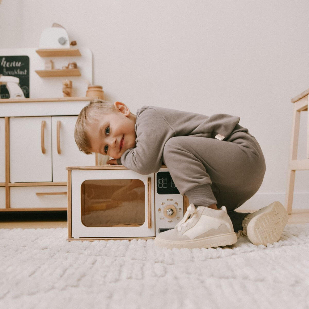 Child playing with a toy microwave in a room with wooden furniture and a white wall.
