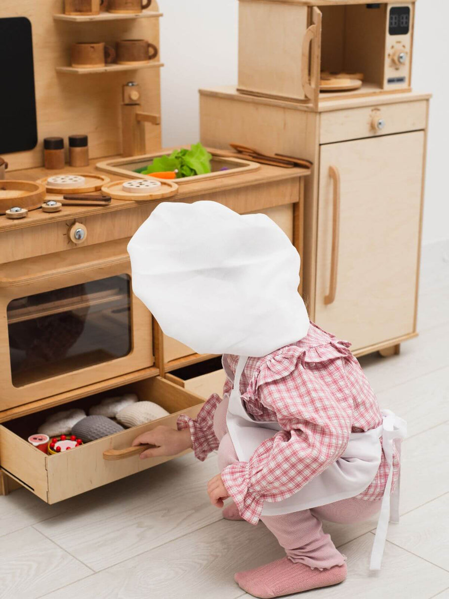 Child in a chef's outfit playing in a wooden play kitchen.
