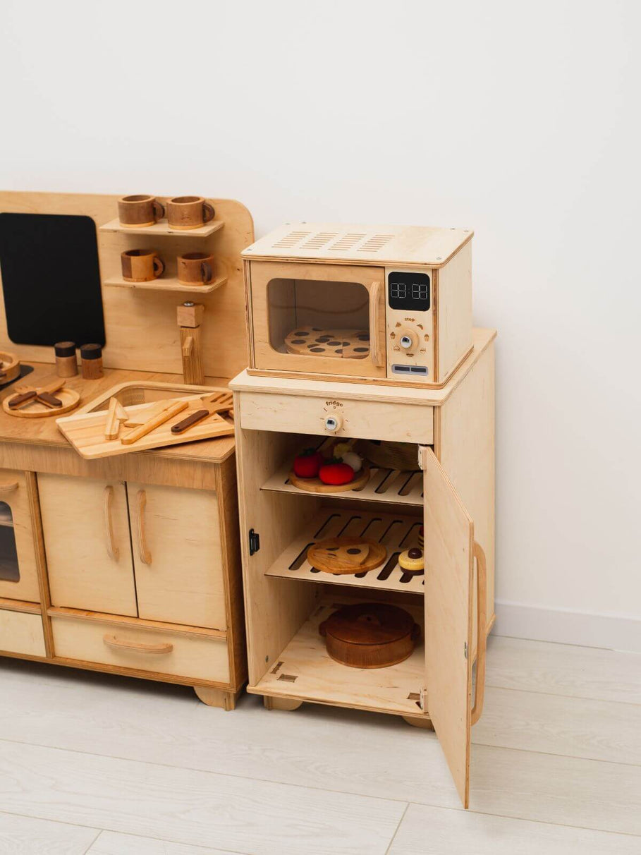 Wooden play kitchen set with various accessories on a white floor and wall.