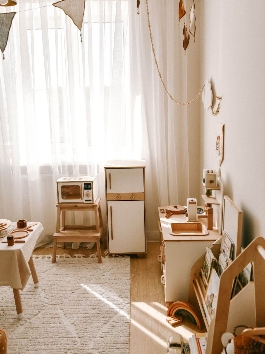 Children's play kitchen set with toy refrigerator, microwave, and sink in a bright room.