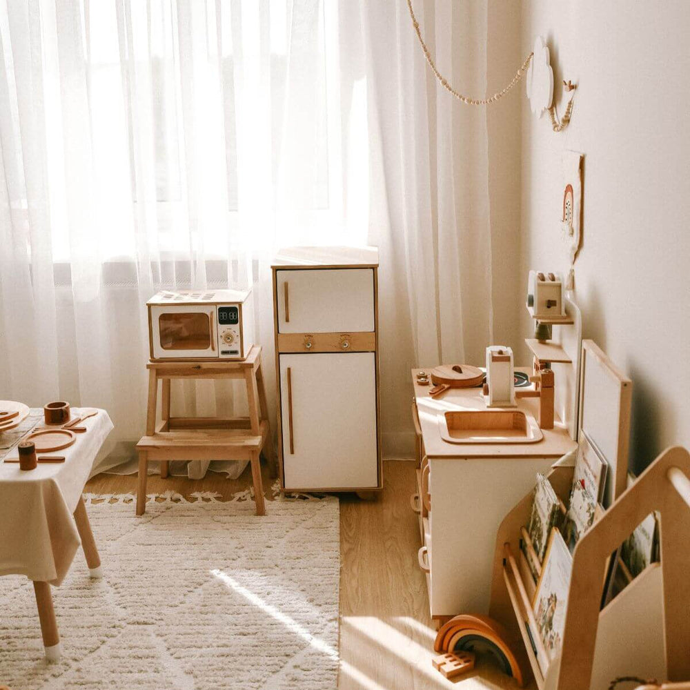 Children's play kitchen set with wooden furniture and toy appliances in a bright room.