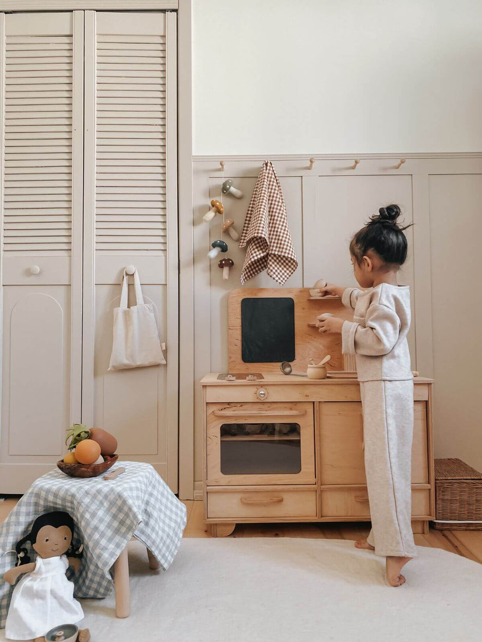 Child playing with a wooden play kitchen set in a room with white walls and wooden furniture.