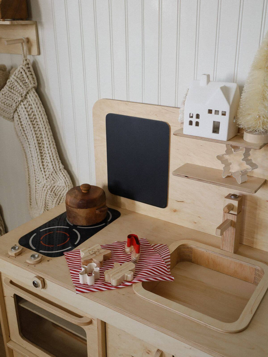 Wooden play kitchen set with stove, sink, and utensils on a white wall background