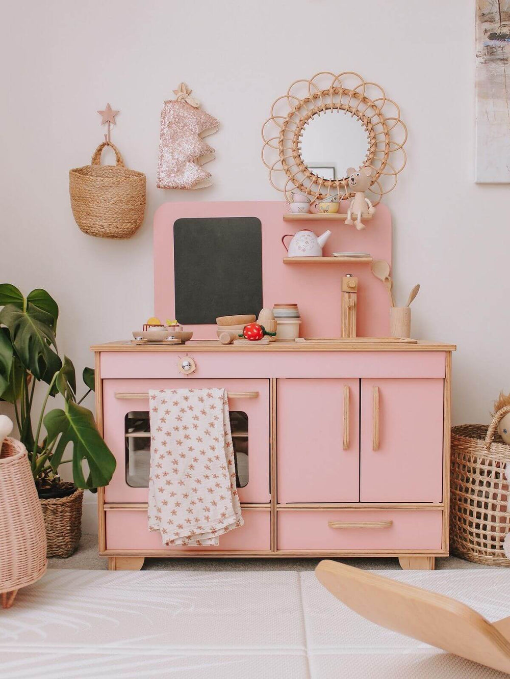 Pink children's play kitchen set with wooden accents in a room with plants and baskets.