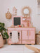 Pink children's play kitchen set with wooden accents in a room with plants and baskets.