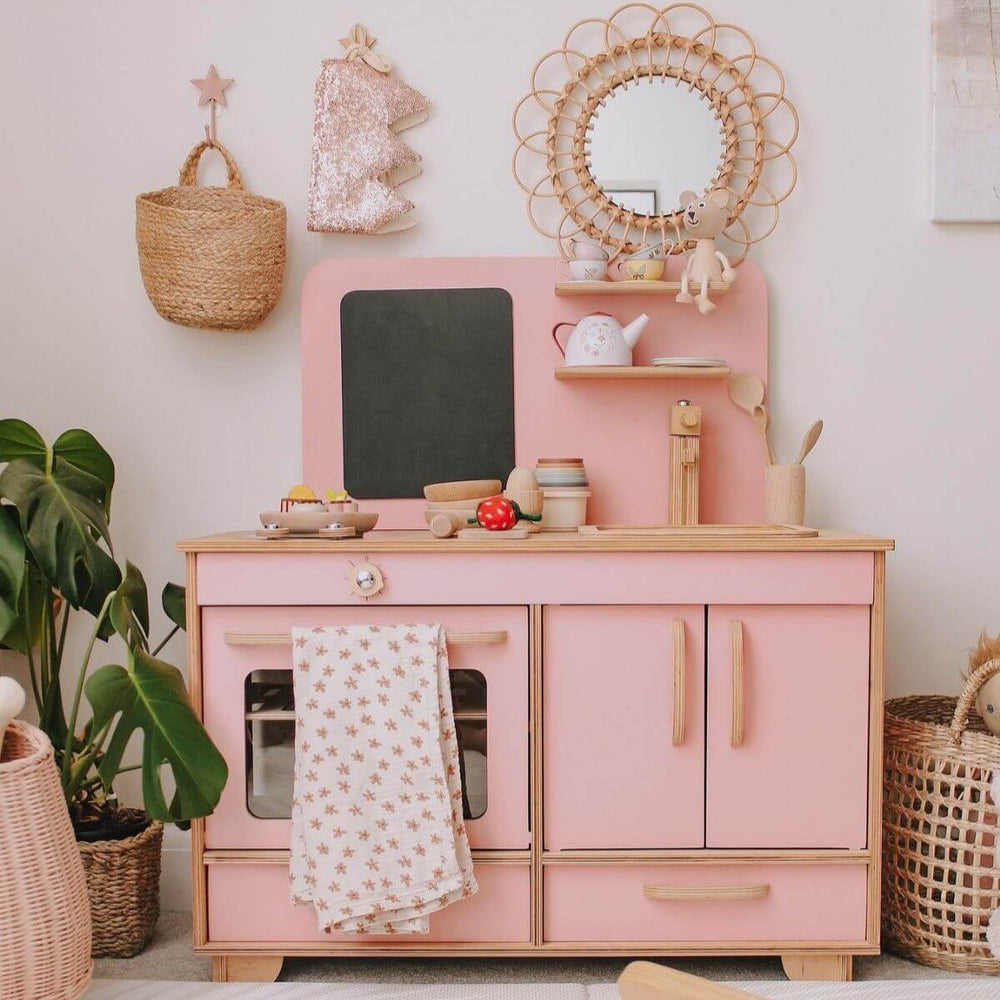 Pink children's play kitchen with wooden accents in a room with plants and decorative items.