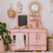 Pink children's play kitchen with wooden accents in a room with plants and decorative items.