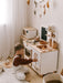 Child playing with a wooden play kitchen set in a room with white curtains and decorative wall art.