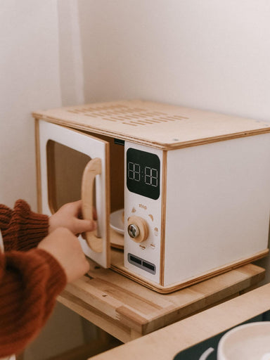 Child opening a white play microwave with wooden frame on a wooden stand.