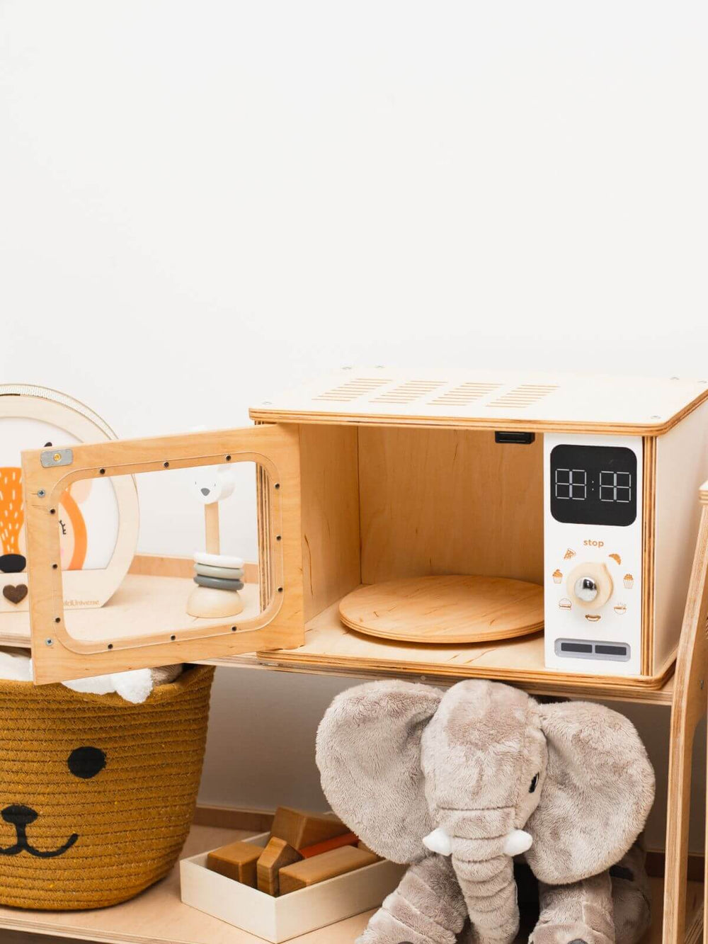 Door open to a white wooden play microwave on a shelf in a playroom with toys surrounding it