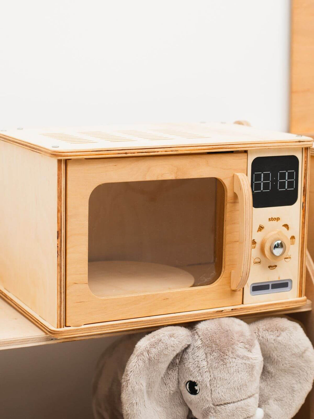 Wooden toy microwave with digital display and control panel, featuring a plush elephant toy below.