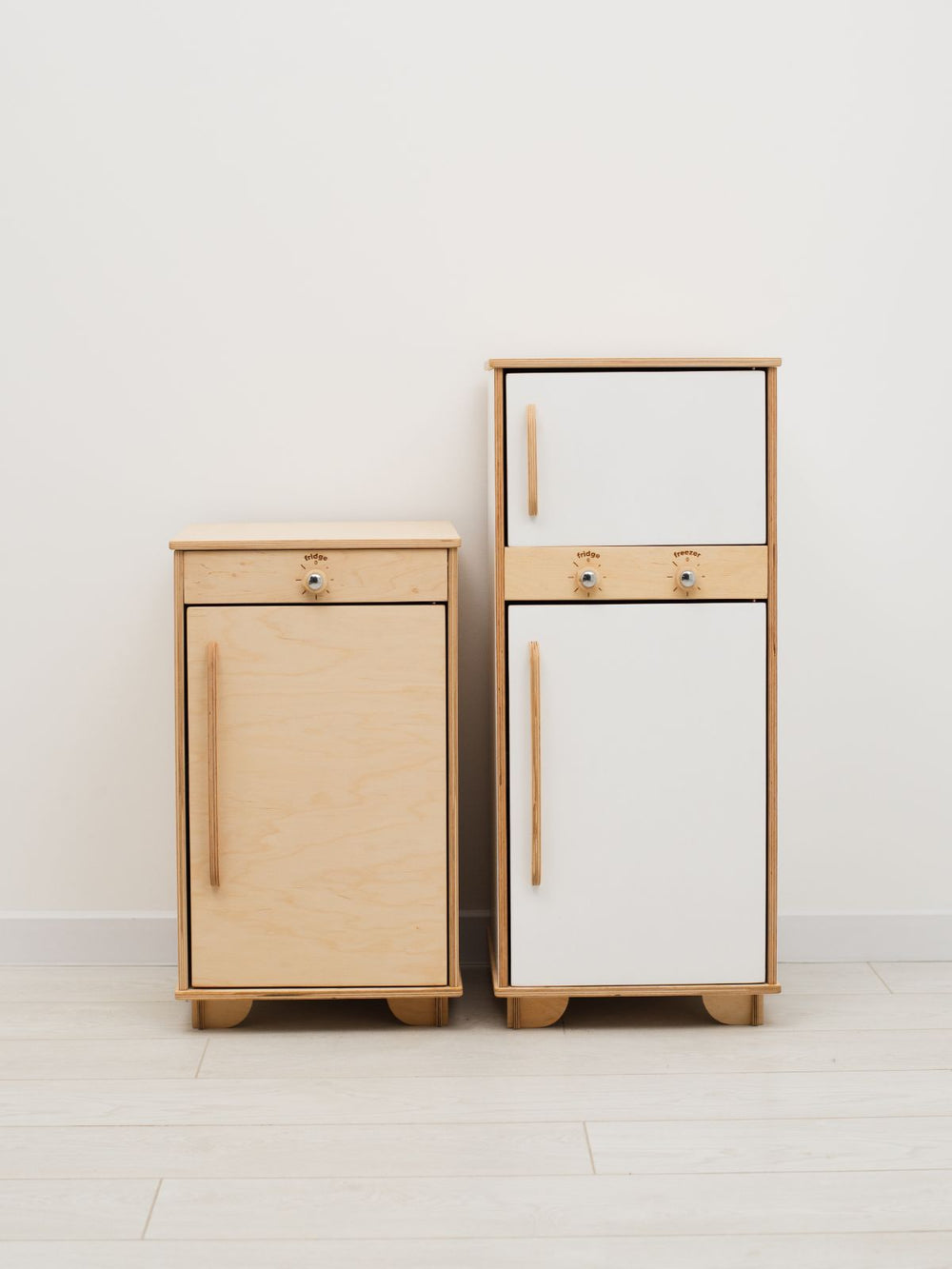 Two wooden play refrigerators, one light wood and one white, on a white floor with a white wall background.