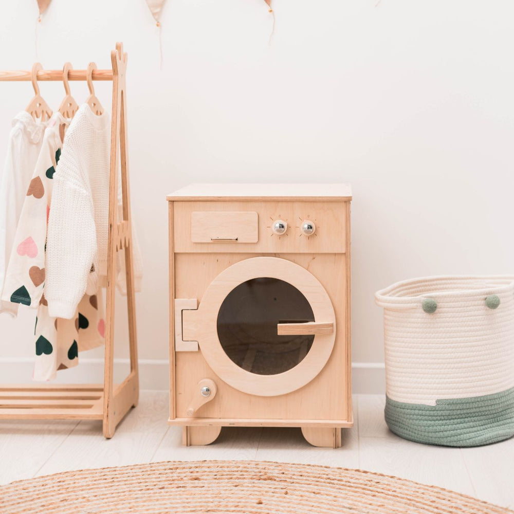 Wooden toy washing machine next to a coat rack with clothes and a basket on a light wooden floor.