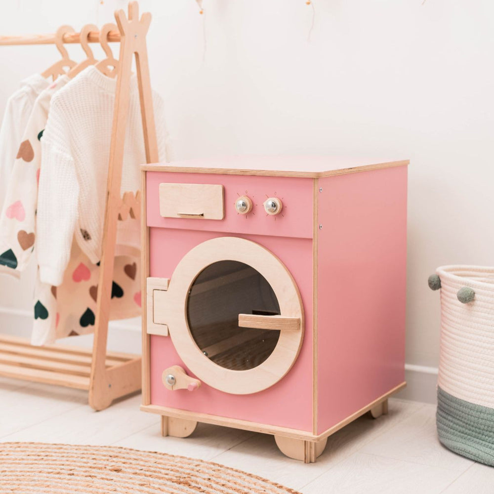 Pink toy washing machine in a child's room with a coat rack and basket in the background.