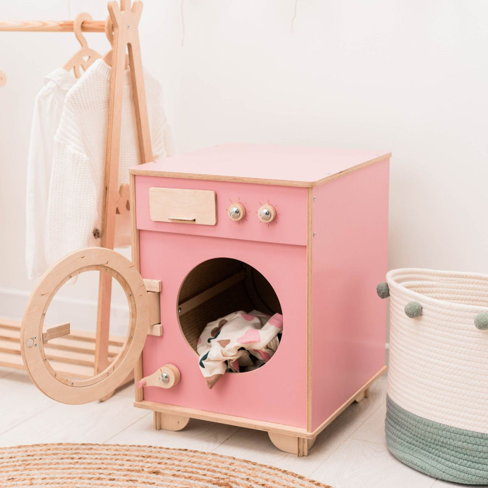 Pink toy washing machine with clothes inside, wooden cart, and basket in a room setting