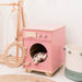 Pink toy washing machine with clothes inside, wooden cart, and basket in a room setting