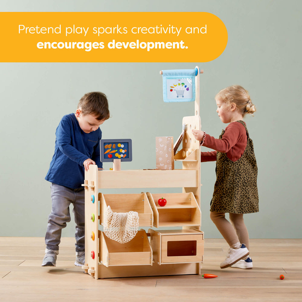 Two children playing with a wooden toy kitchen set in a room with educational posters on the wall.