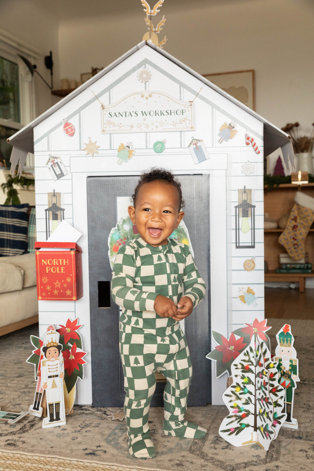 Child in a checkered outfit standing in front of a festive playhouse with Christmas decorations.