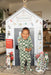 Child in a checkered outfit standing in front of a festive playhouse with Christmas decorations.