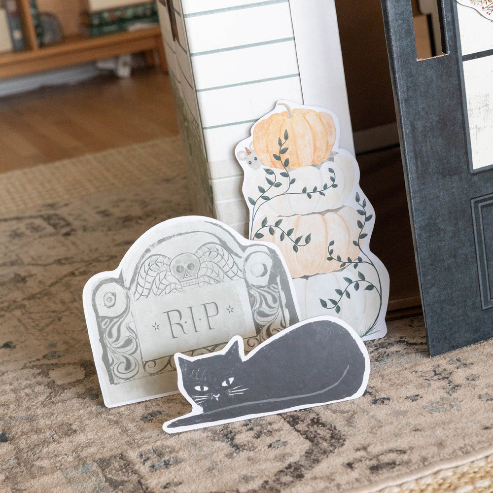 Decorative Halloween items including a gravestone, pumpkins, and a cat in front of a playhouse.
