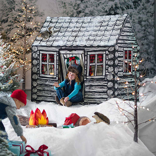 Children playing around a play log cabin in a snowy scene with Christmas decorations.