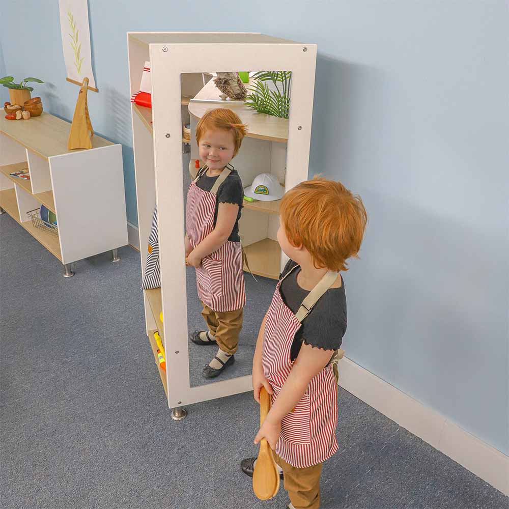 Child wearing an apron looking at a mirror on a dress-up wardrobe in a room with shelves and plants.