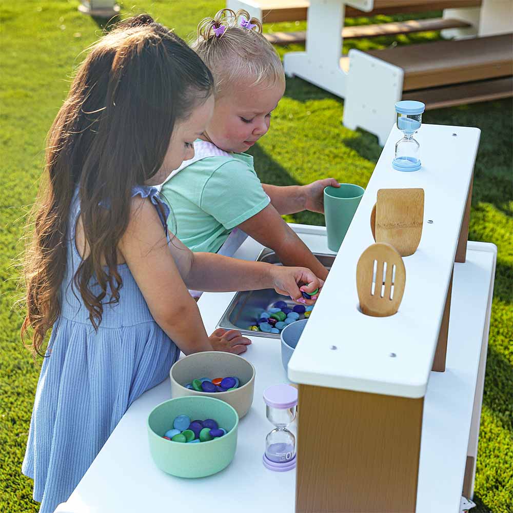 Two young girls playing with an outdoor mud kitchen set on grass.