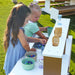 Two young girls playing with an outdoor mud kitchen set on grass.