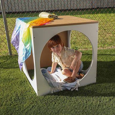 Child playing inside an outdoor play cube with a colorful towel draped over it.