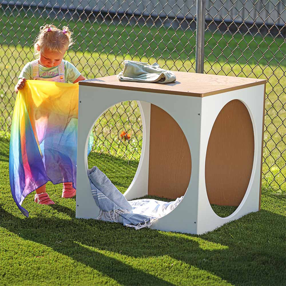 Child playing with a colorful blanket near a wooden outdoor play cube.
